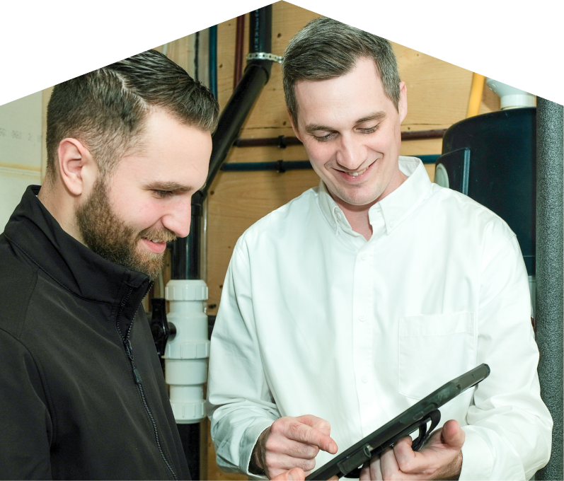 Haven Home specialist reviewing maintenance plan options with a smiling homeowner using a tablet, inside a utility room with visible piping. Haven Home specialist reviewing maintenance plan options with a smiling homeowner using a tablet, inside a utility room with visible piping.