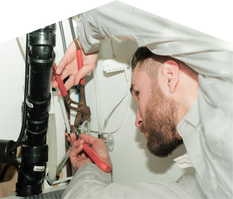 Haven Home plumbing specialist uses hand tools to tighten pipe fittings beneath a sink during a repair, with visible wiring and drainage lines in the background. Haven Home plumbing specialist uses hand tools to tighten pipe fittings beneath a sink during a repair, with visible wiring and drainage lines in the background.