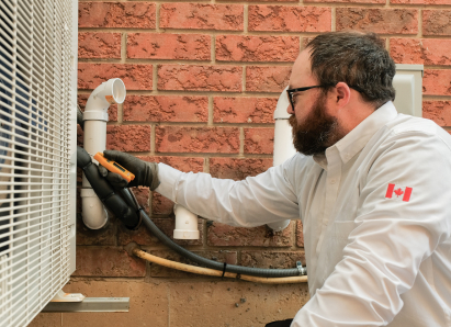 A Haven Home HVAC specialist wearing glasses and gloves uses a diagnostic tool to service an outdoor AC unit beside a brick wall. A Haven Home HVAC specialist wearing glasses and gloves uses a diagnostic tool to service an outdoor AC unit beside a brick wall.